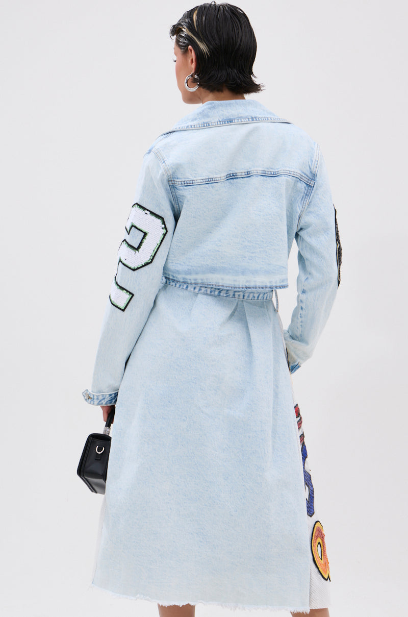 A person with short dark hair, large hoop earrings, and a small black handbag is seen from behind in the COUNTDOWN PATCH DENIM TRENCH—a long light blue coat with varsity-style patches—for a sporty-chic look. 4