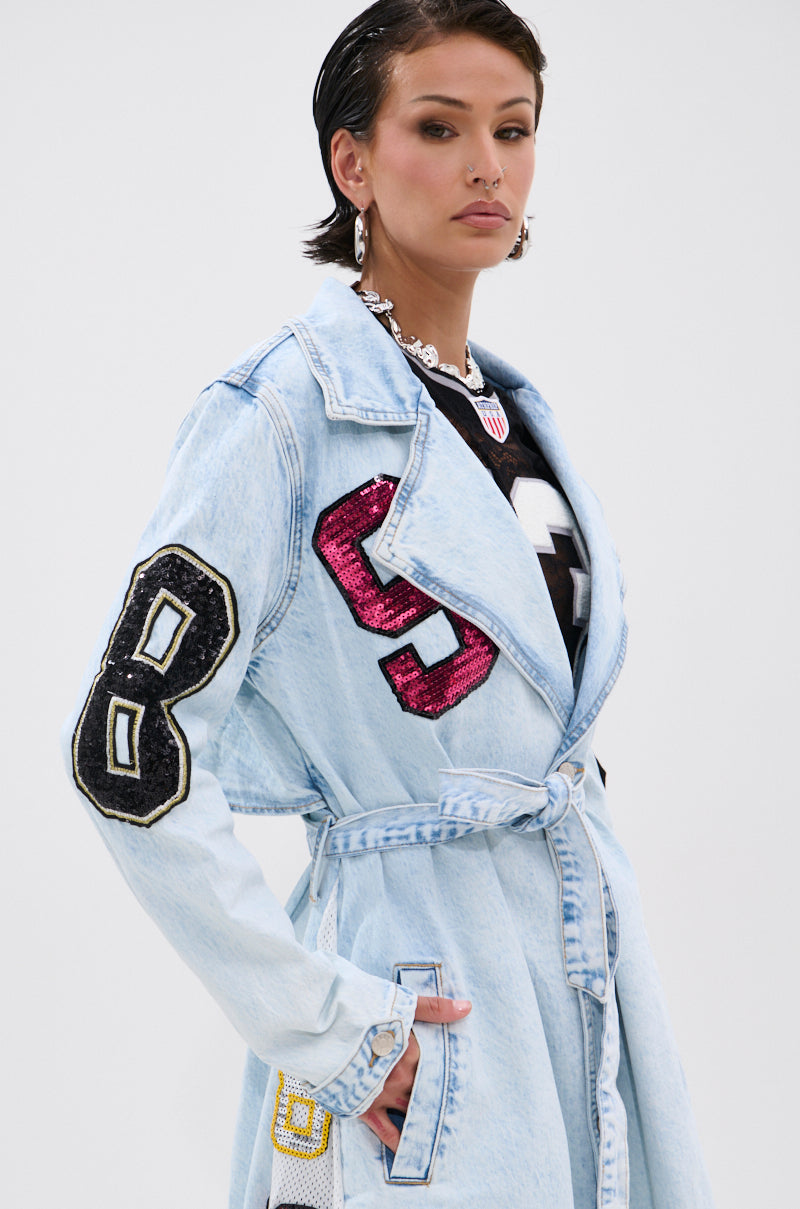 Wearing the COUNTDOWN PATCH DENIM TRENCH—a light blue denim coat with sporty varsity-style patches—someone stands confidently, accessorized with hoop earrings and a headband, against a plain background. 6