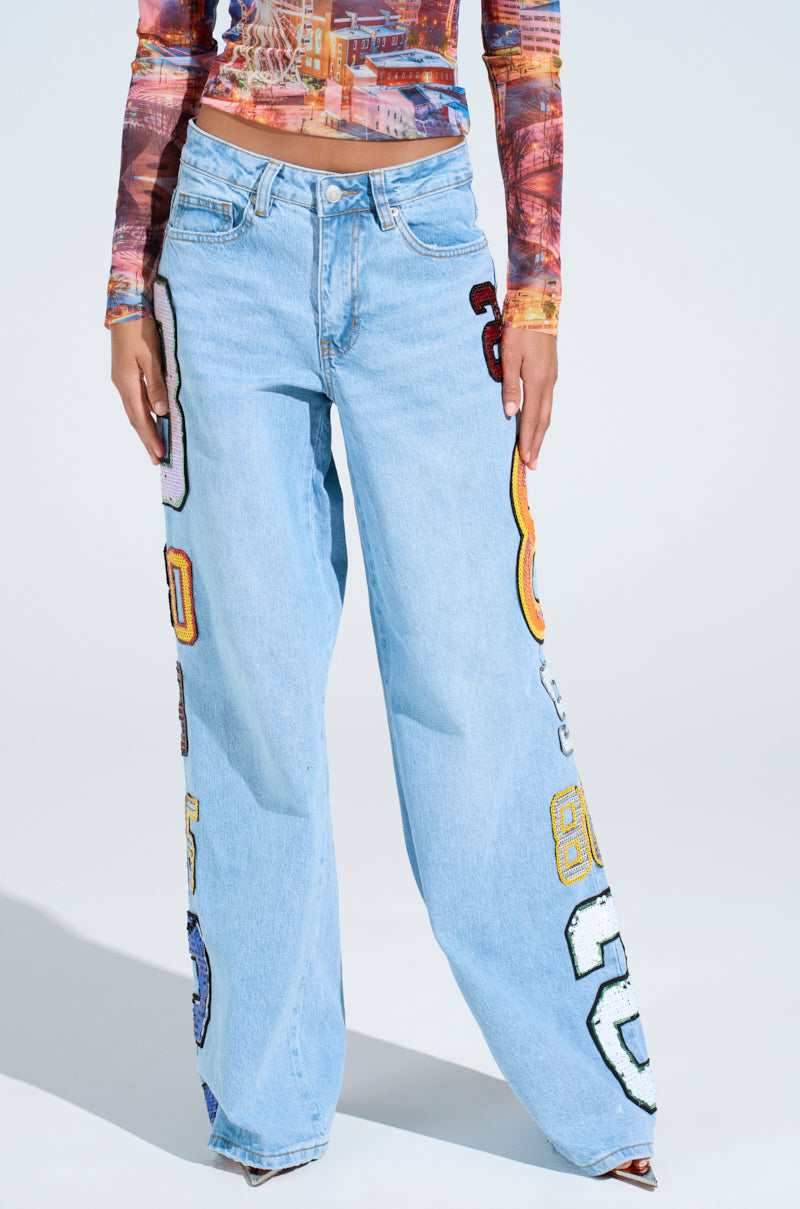 A person wearing the GOOD LUCK PATCH DENIM—high-rise, light blue wide-leg jeans with oversized sequin patches and colorful number appliqués—paired with a long-sleeve printed fitted top, stands against a white background.