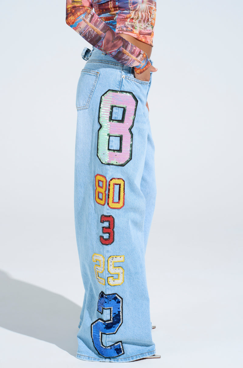 Wearing the GOOD LUCK PATCH DENIM—high-rise, light blue wide-leg jeans with bold, colorful number patches—paired with a vibrant long-sleeved top.