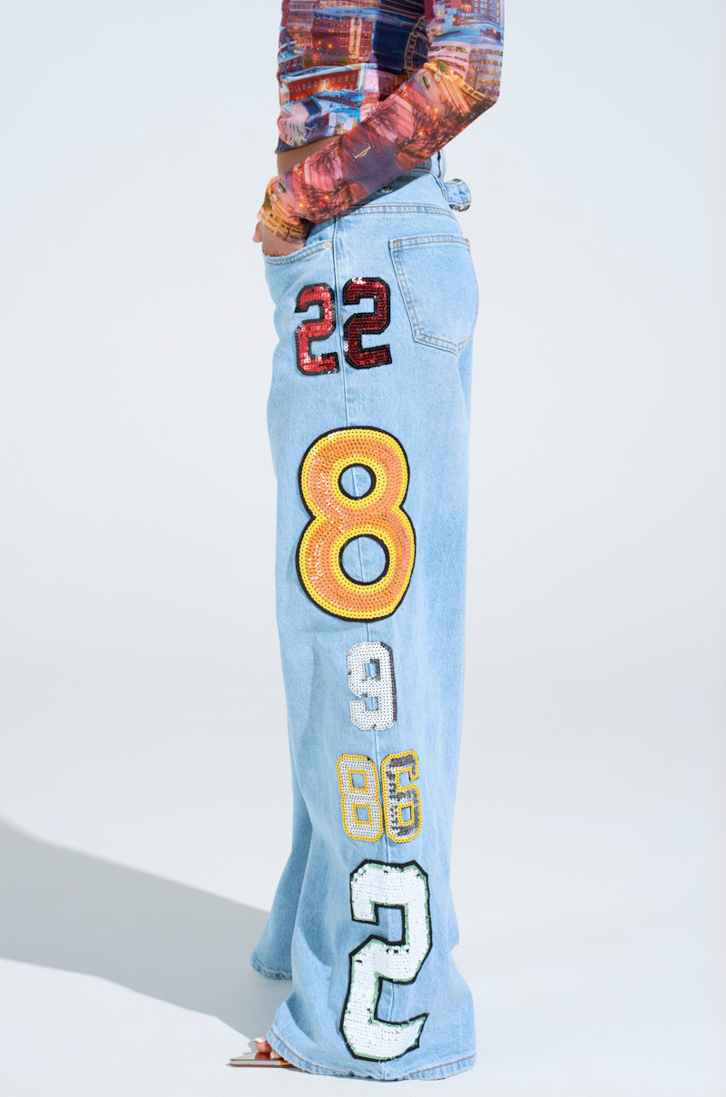 Someone sports the GOOD LUCK PATCH DENIM, high-rise wide-leg jeans featuring bold, colorful embroidered numbers down one leg, styled with a patterned long-sleeve top for a playful statement.
