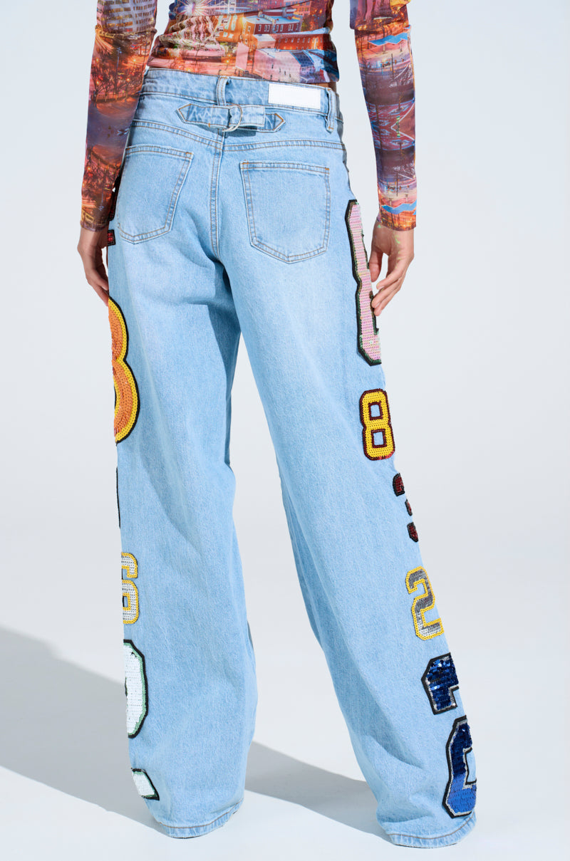 Model wearing GOOD LUCK PATCH DENIM high-rise wide-leg jeans with oversized sequin number patches on the legs and back pockets, paired with a multicolored long-sleeve top, shown from the back against a light background.