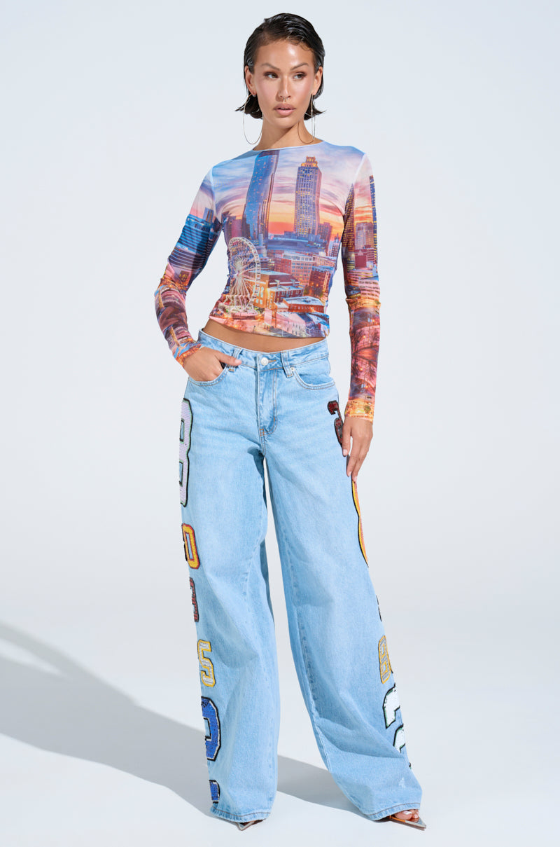 A woman models the GOOD LUCK PATCH DENIM—high-rise, wide-leg jeans with oversized sequin patches—and a cityscape-print long-sleeve top, posing confidently against a plain white background.