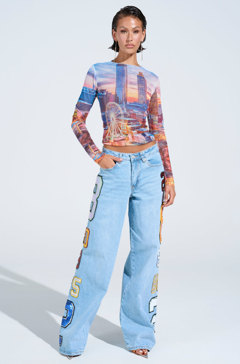 A woman stands confidently in the GOOD LUCK PATCH DENIM—high-rise, wide-leg jeans with large, colorful numbers—paired with a cityscape print long-sleeve top and open-toe heels against a plain white background.