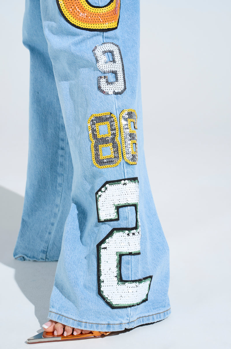 A person wears the GOOD LUCK PATCH DENIM, light blue high-rise jeans with a wide-leg fit and oversized sequin patches of 9, 86, and 2, paired with brown sandals against a plain light background.