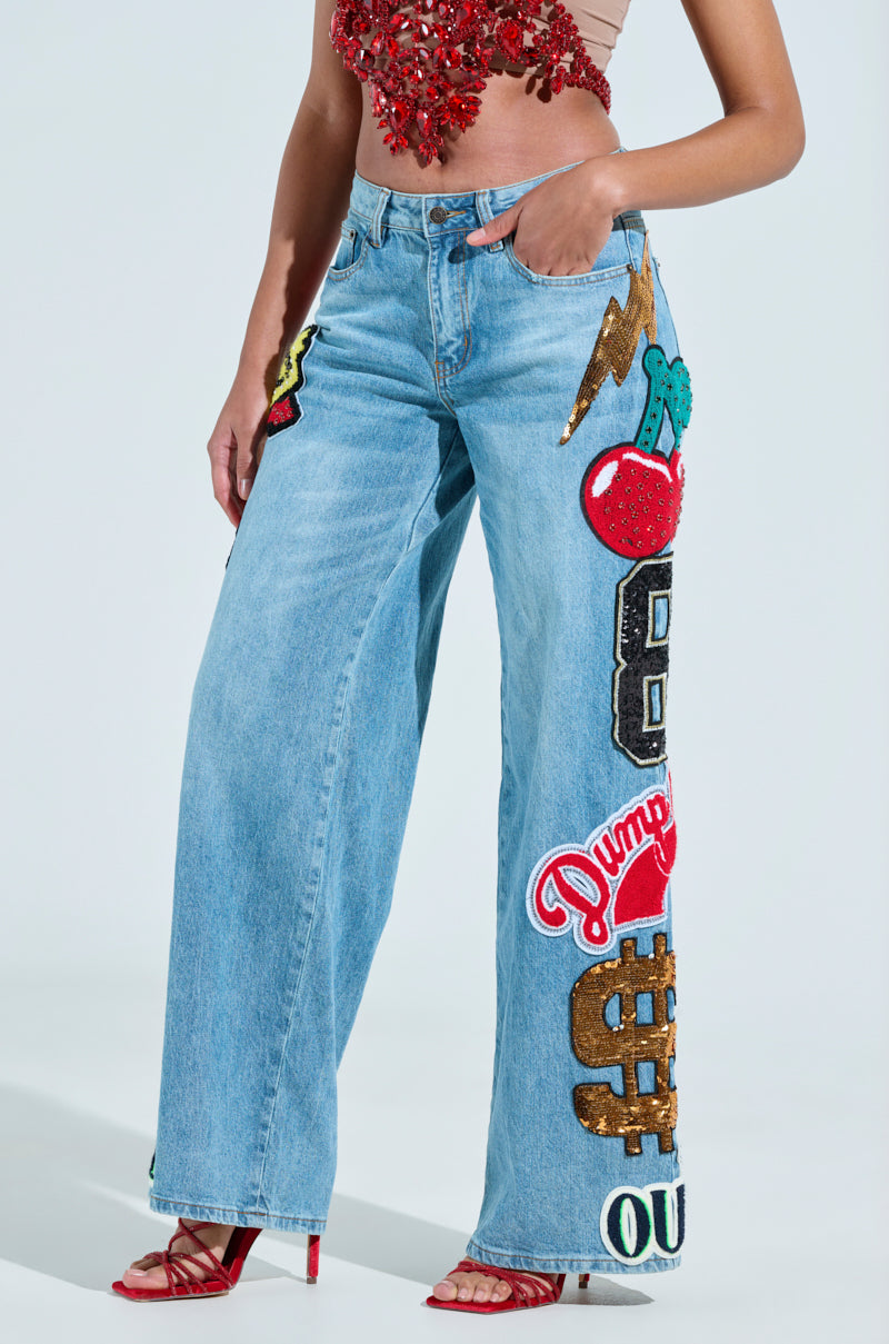 A person models the CHECK IT OUT OVERSIZED PATCH JEANS, featuring vibrant denim patches like a lightning bolt, cherries, and various letters. They're styled with a red beaded crop top and red strappy heels.