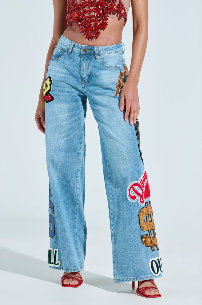 Wearing CHECK IT OUT OVERSIZED PATCH JEANS with vibrant denim patches featuring dollar signs and text, they pair it with a textured red top and red strapped heels, posing against a plain background.