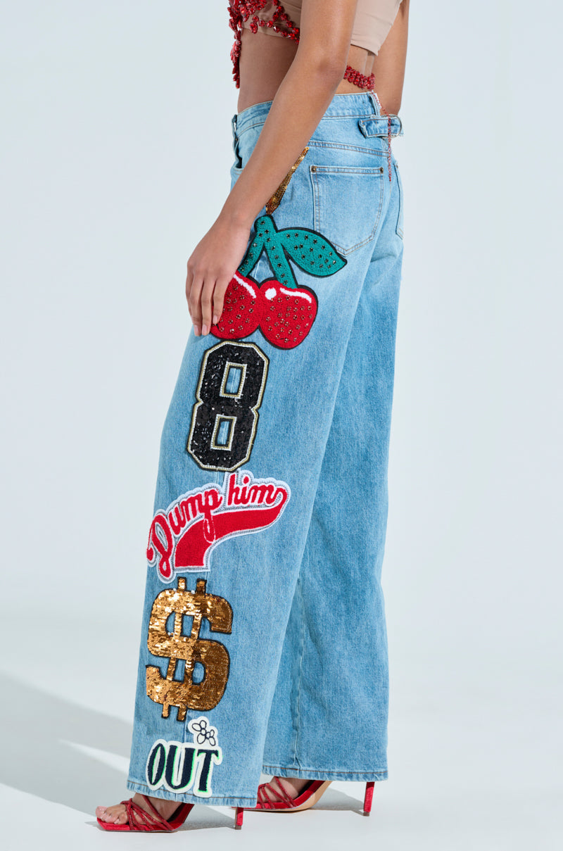 A person wears the Check It Out Oversized Patch Jeans, featuring light blue, high-rise wide-leg style with vibrant patches like cherries, the number 8, and phrases "Dump him" and "$ OUT." To complete the look, they pair it with a sleeveless top and red strappy sandals.