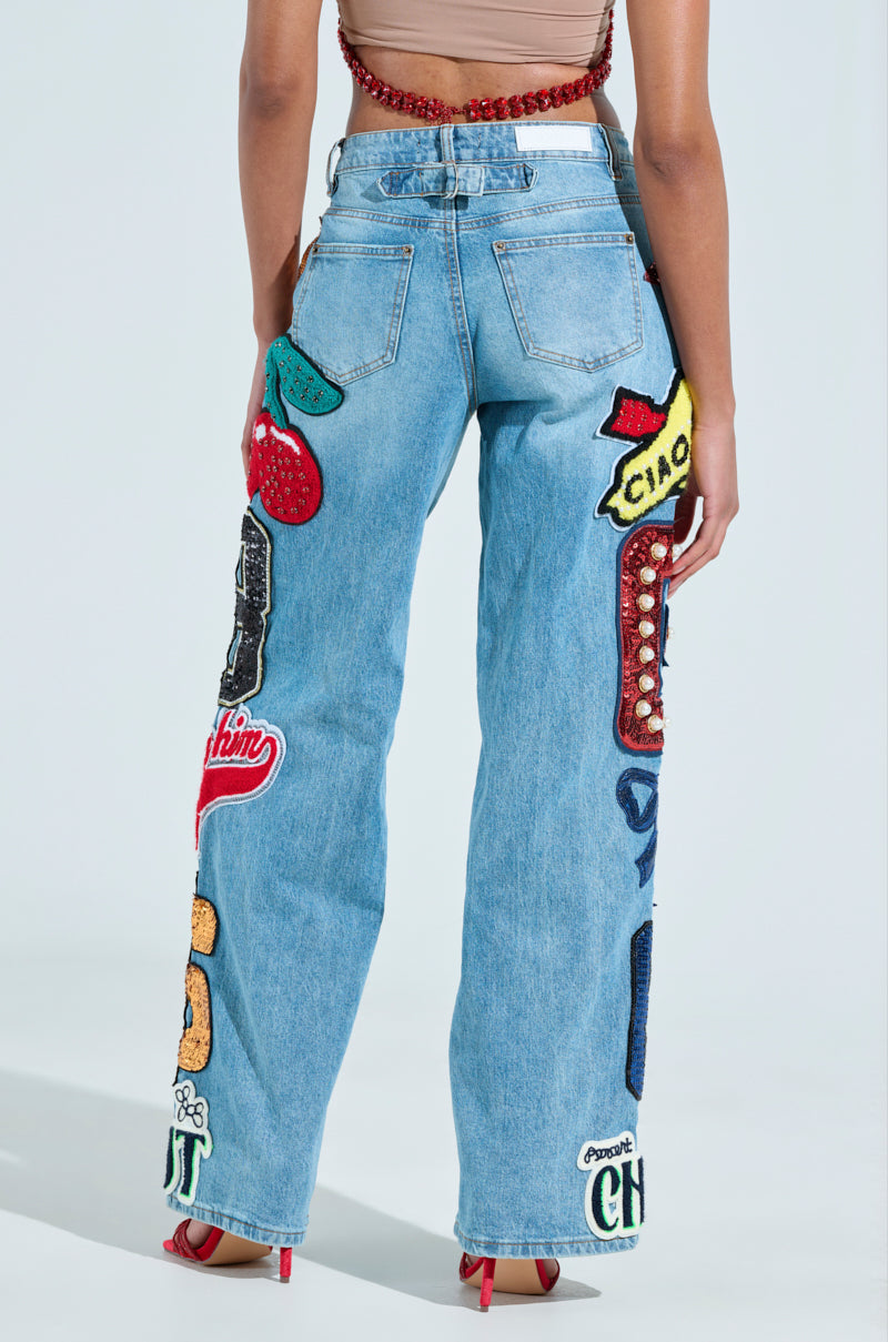 Rear view of a person wearing CHECK IT OUT OVERSIZED PATCH JEANS in light blue with vibrant denim patches, paired with red heeled sandals and a beige top against a plain white background.