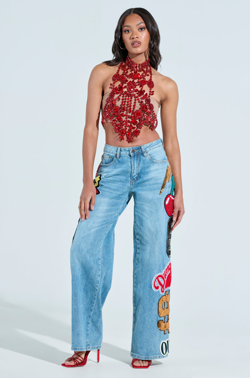 A woman poses in a red intricately designed halter top paired with CHECK IT OUT OVERSIZED PATCH JEANS, featuring vibrant patches and graphics. She completes her look with red high-heeled sandals and loose waves in her hair.