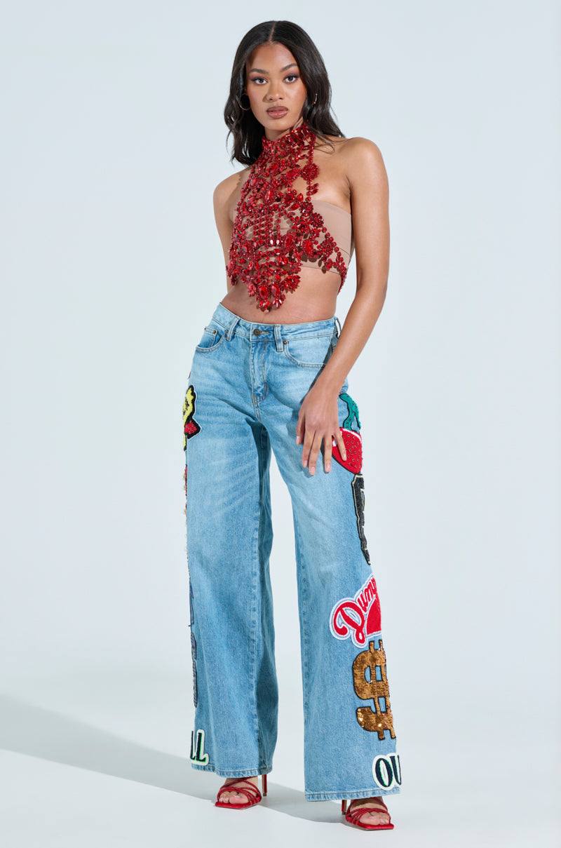 A woman confidently models in a red crochet halter top paired with CHECK IT OUT OVERSIZED PATCH JEANS, featuring vivid denim patches. She sports red sandals and stands against a light, neutral background, exuding effortless style.