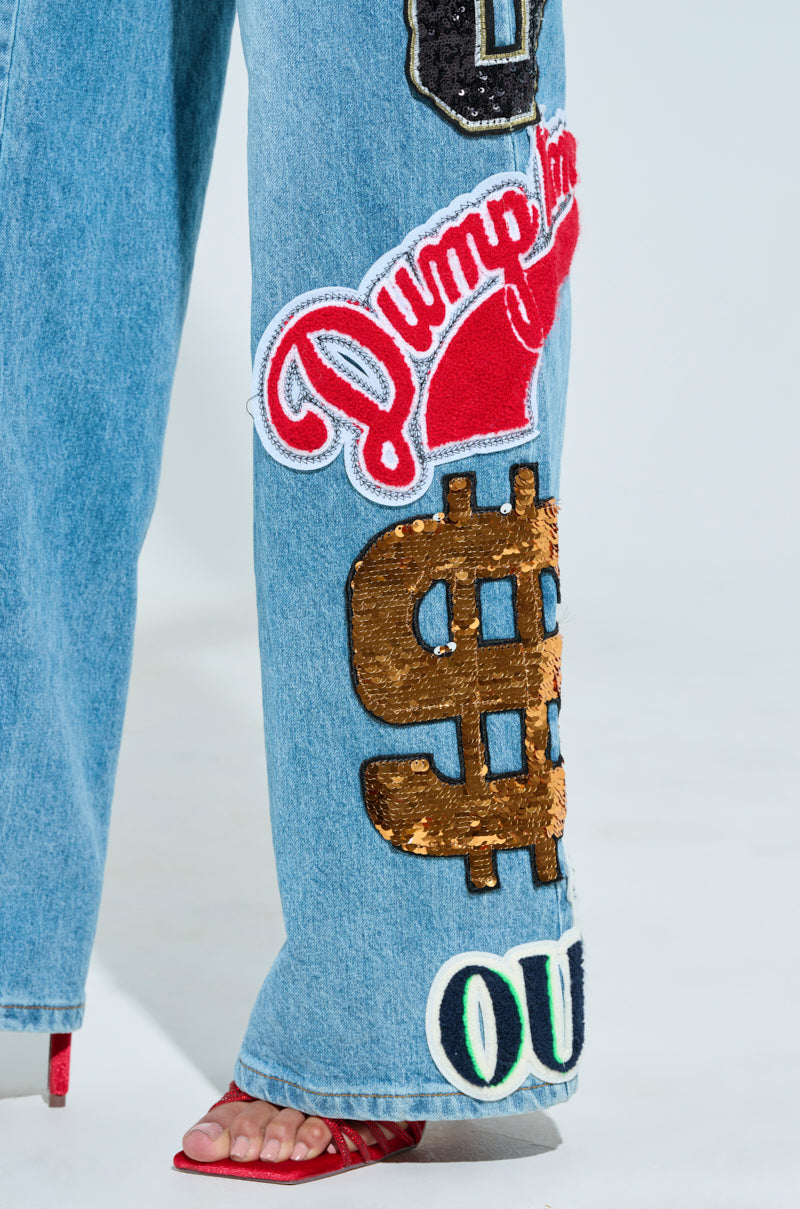Against a light blue backdrop, the CHECK IT OUT OVERSIZED PATCH JEANS are showcased with close-up details of high-rise wide-leg denim featuring vibrant oversized patches like "DUMP HIM" and a sequined gold dollar sign. Paired with red strappy sandals for an eye-catching look.