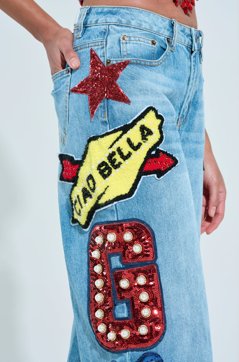 A person in CHECK IT OUT OVERSIZED PATCH JEANS, featuring a red star, "CIAO BELLA" yellow patch, and letterman-style red "G" with pearls, casually slips a hand into the front pocket.