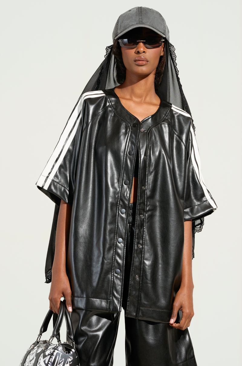 A person exudes sporty-chic style wearing the EXTRA BUTTER LEATHER BUTTON DOWN shirt and pants in black. Their look is completed with a gray cap, sunglasses, a headscarf, and a patterned bag in one hand, achieving an edgy and contemporary vibe.