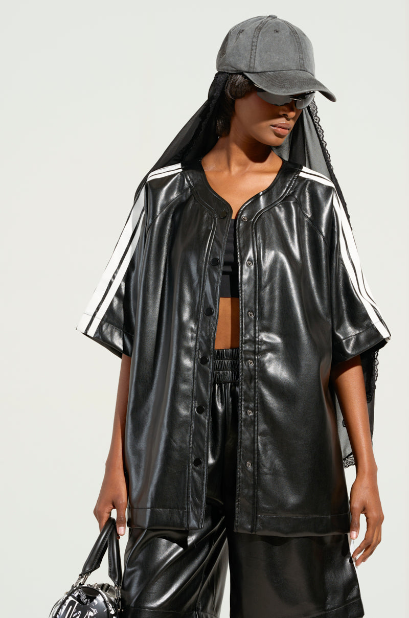 A person showcasing a sporty-chic vibe in the EXTRA BUTTER LEATHER BUTTON DOWN, featuring a loose-fitting faux leather design with white stripes. They pair it with matching leather pants and accessorize with a gray cap adorned with a black veil. Holding a patterned handbag, they stand against a plain background.