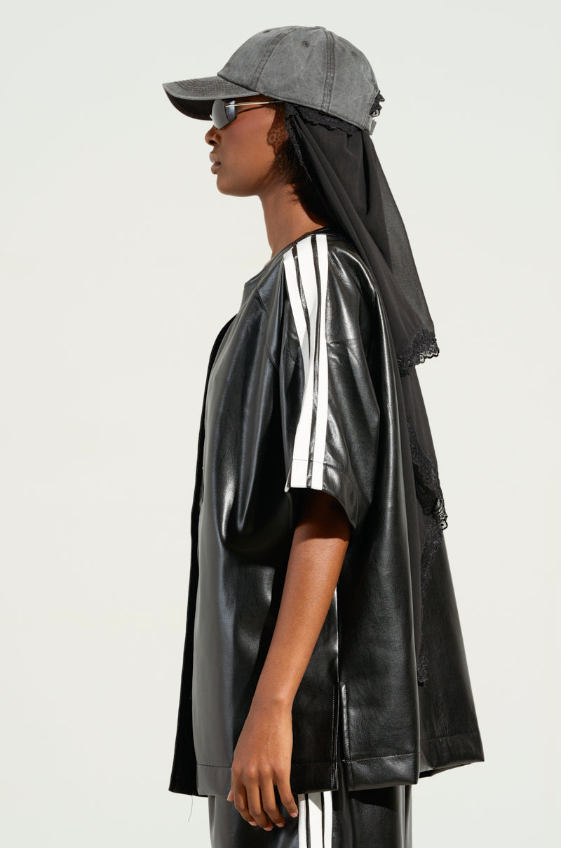 A person stands in profile, exuding sporty-chic vibes in an EXTRA BUTTER LEATHER BUTTON DOWN with white stripes. A dark baseball cap and sunglasses enhance the outfit, while a sheer black fabric cascades elegantly over the back of the cap.