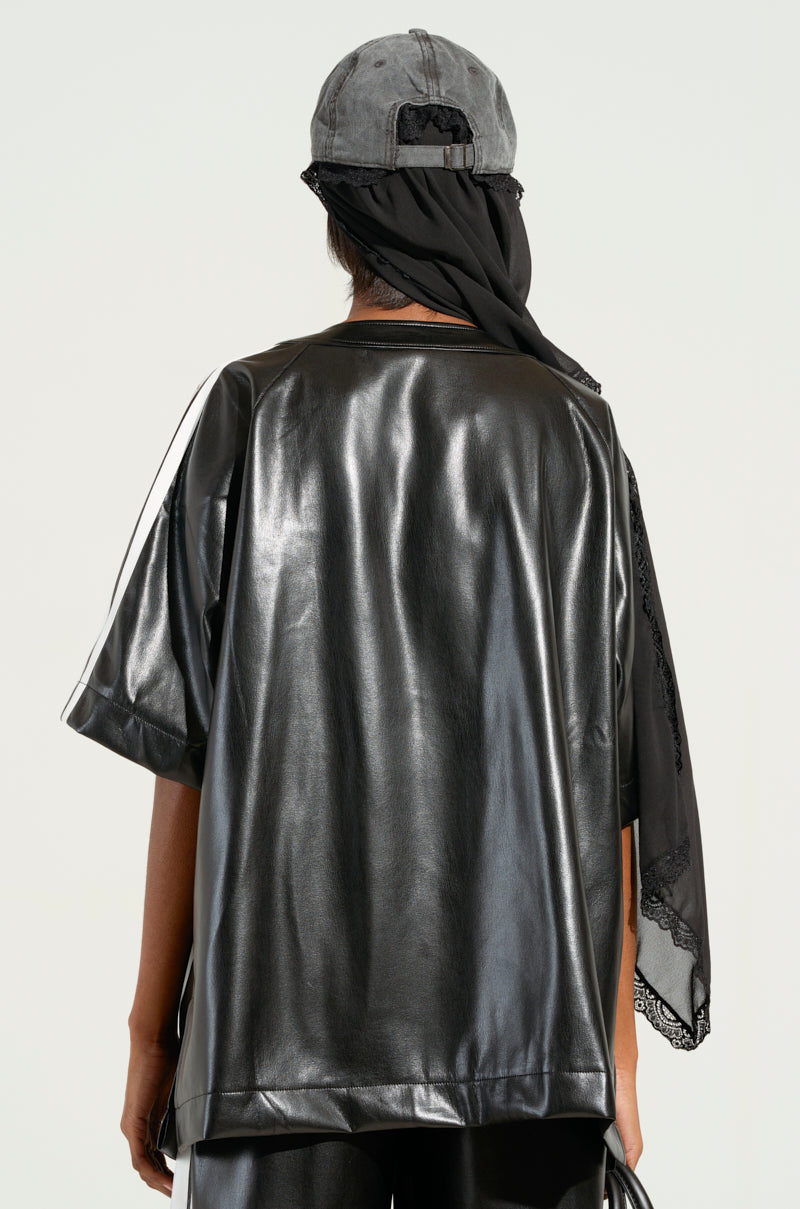 A person wearing an EXTRA BUTTER LEATHER BUTTON DOWN and a gray baseball cap stands facing away. A black scarf drapes over the cap, partially covering the shoulder. The plain light background enhances their sporty-chic vibe.