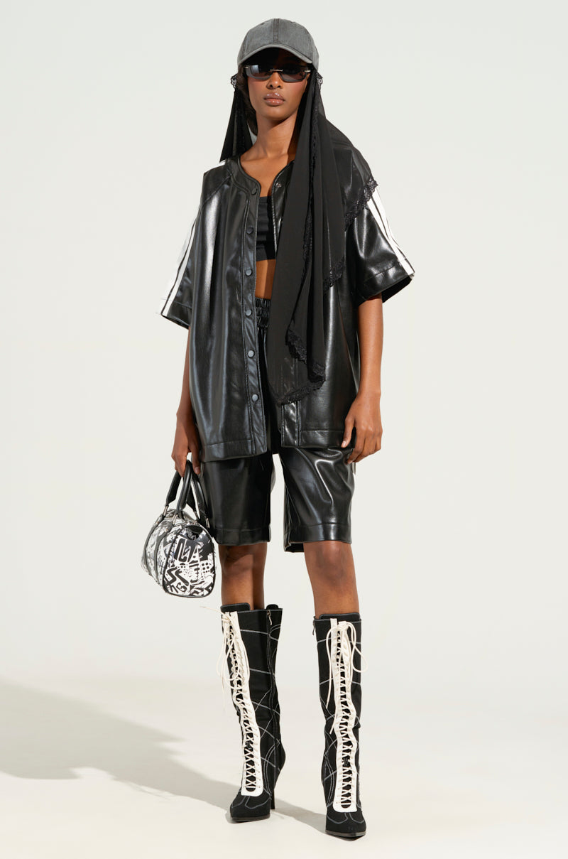 A person wearing the sporty-chic black EXTRA BUTTER LEATHER BUTTON DOWN paired with matching shorts. They accessorize with a gray cap, dark sunglasses, and a patterned bag. Knee-high boots featuring a white lace design complete the look.