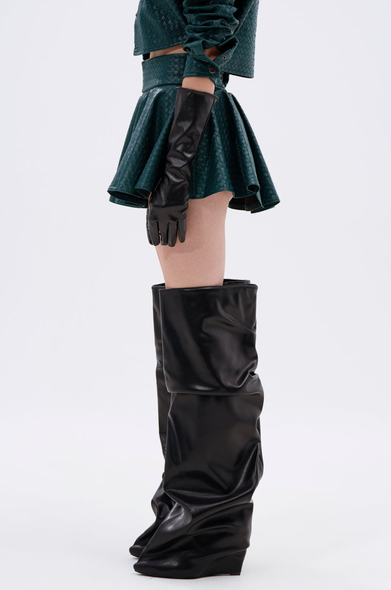 A person stands sideways against a plain light background, wearing the TOO GOOD FOR YOU GREEN MINI SKIRT with a matching textured green crop top, long black gloves, and slouchy over-the-knee black boots. 4