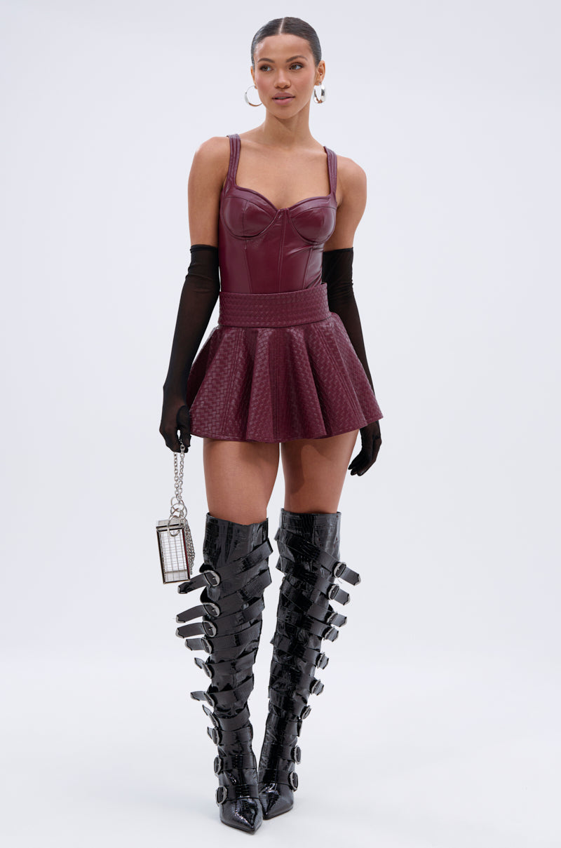 A woman wears the TOO GOOD FOR YOU MINI SKIRT in burgundy faux leather with a fitted bodice and flared hem, paired with long black gloves, tall shiny buckle boots, a small silver clutch, and her hair tied back. 0