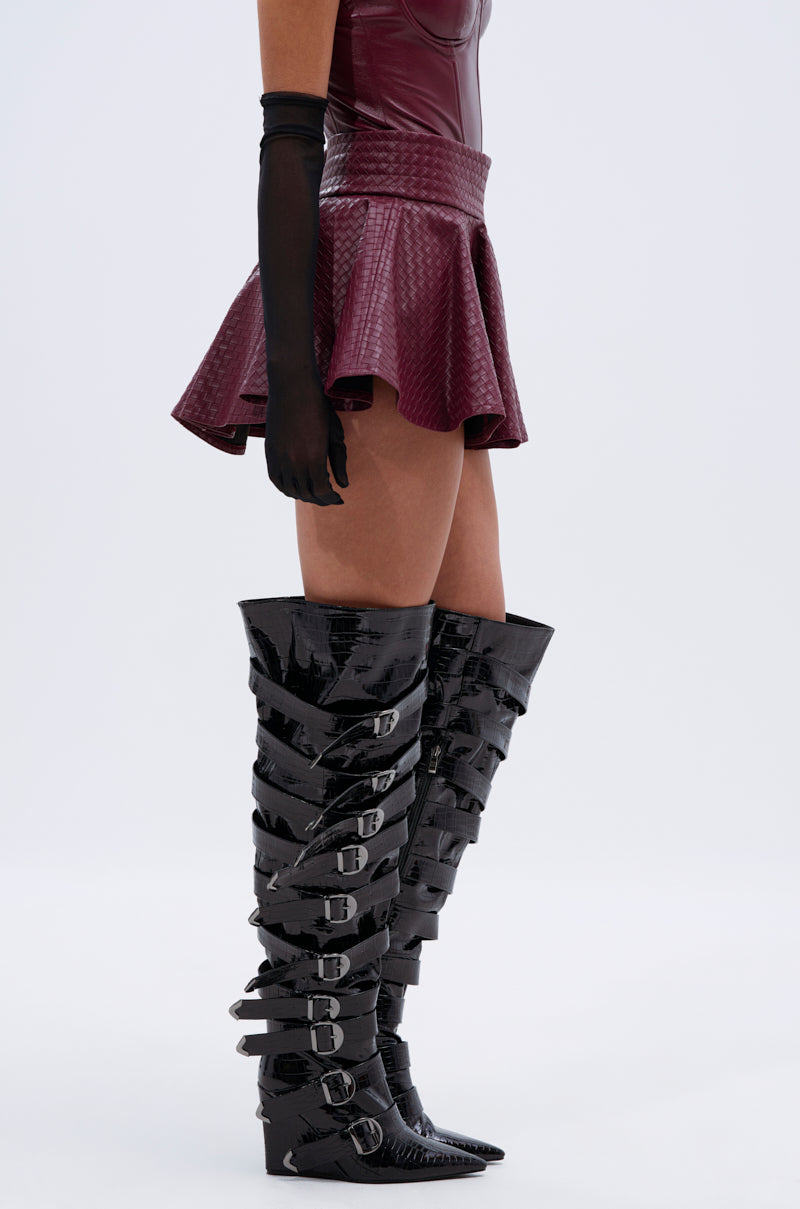 Wearing the TOO GOOD FOR YOU MINI SKIRT in burgundy faux leather with a matching sleeveless top, long black gloves, and black patent knee-high boots with silver buckles, a person stands against a plain light background. 3