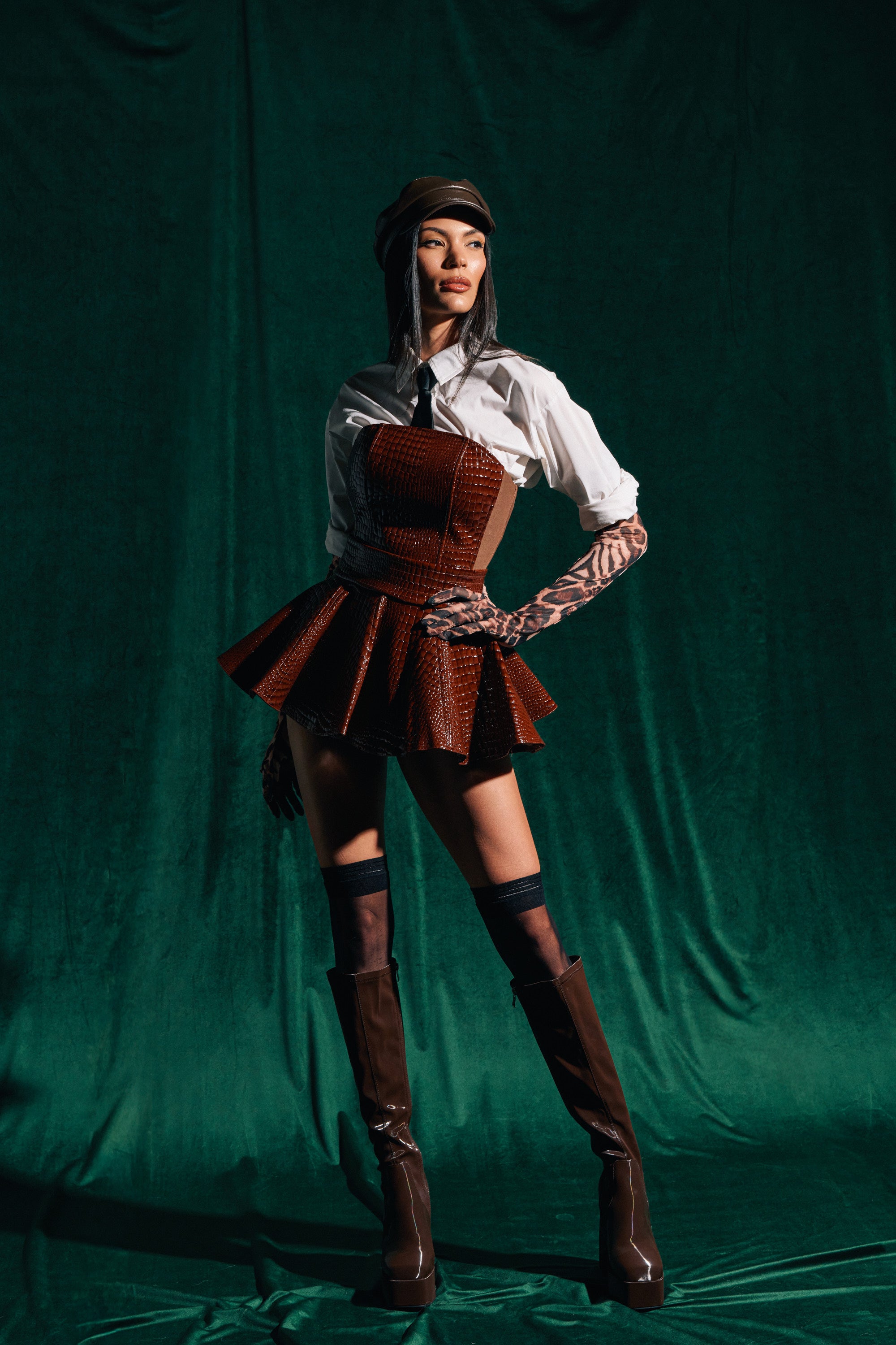 A person poses confidently in a stylish outfit with a white shirt, patterned gloves, high brown boots, a matching beret, and the TOO GOOD FOR YOU CROCODILE MINI SKIRT against a dark green fabric backdrop.