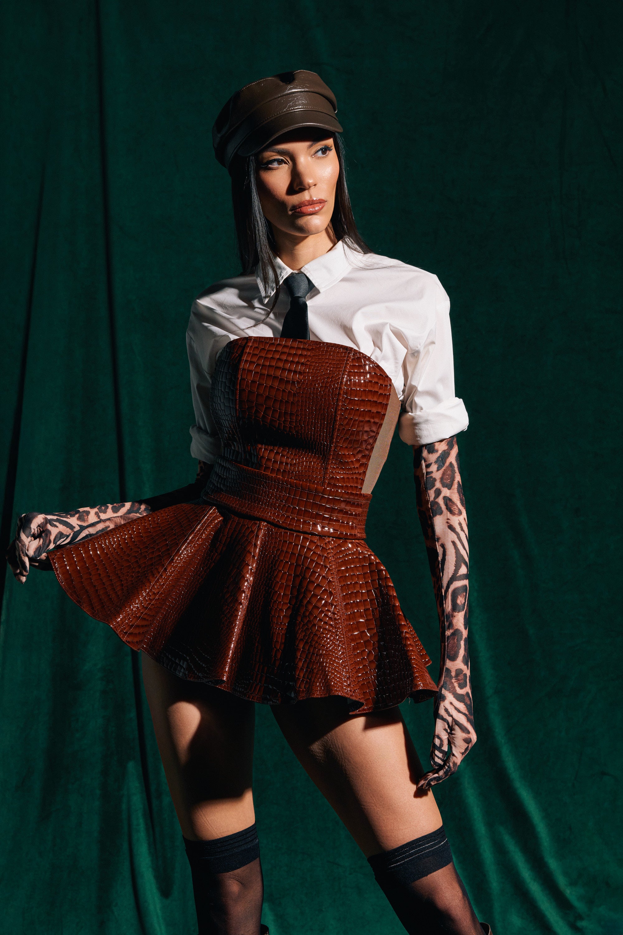 A woman poses against a dark green background in a brown leather cap, white shirt, black tie, and the TOO GOOD FOR YOU CROCODILE MINI SKIRT with a flared silhouette, finishing her look with sheer leopard print gloves.