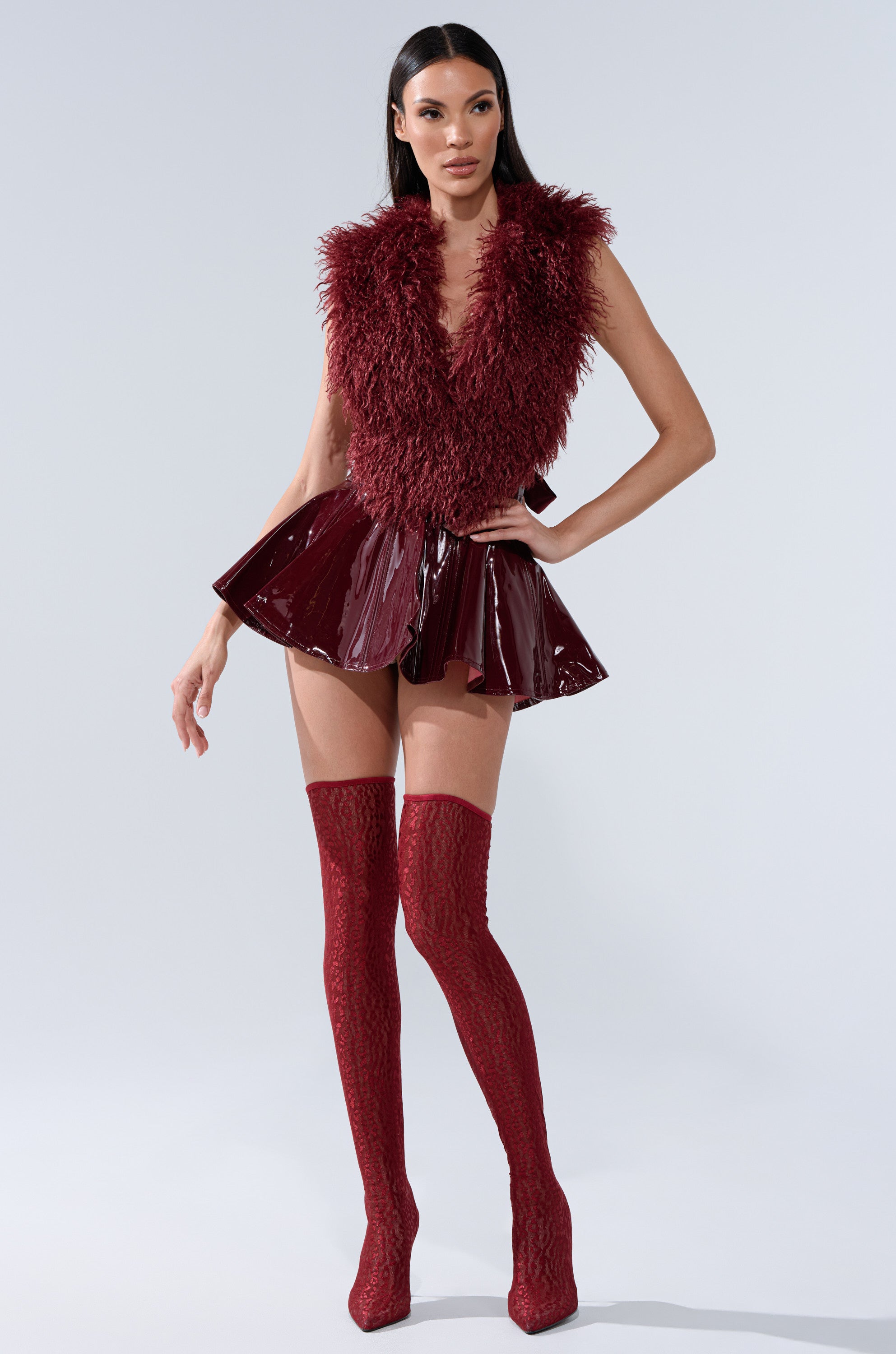 A woman models the SHE IS ON FIRE FAUX LEATHER MINI SKIRT IN BURGUNDY, paired with a burgundy shaggy fur vest and matching thigh-high textured boots, set against a plain light background.