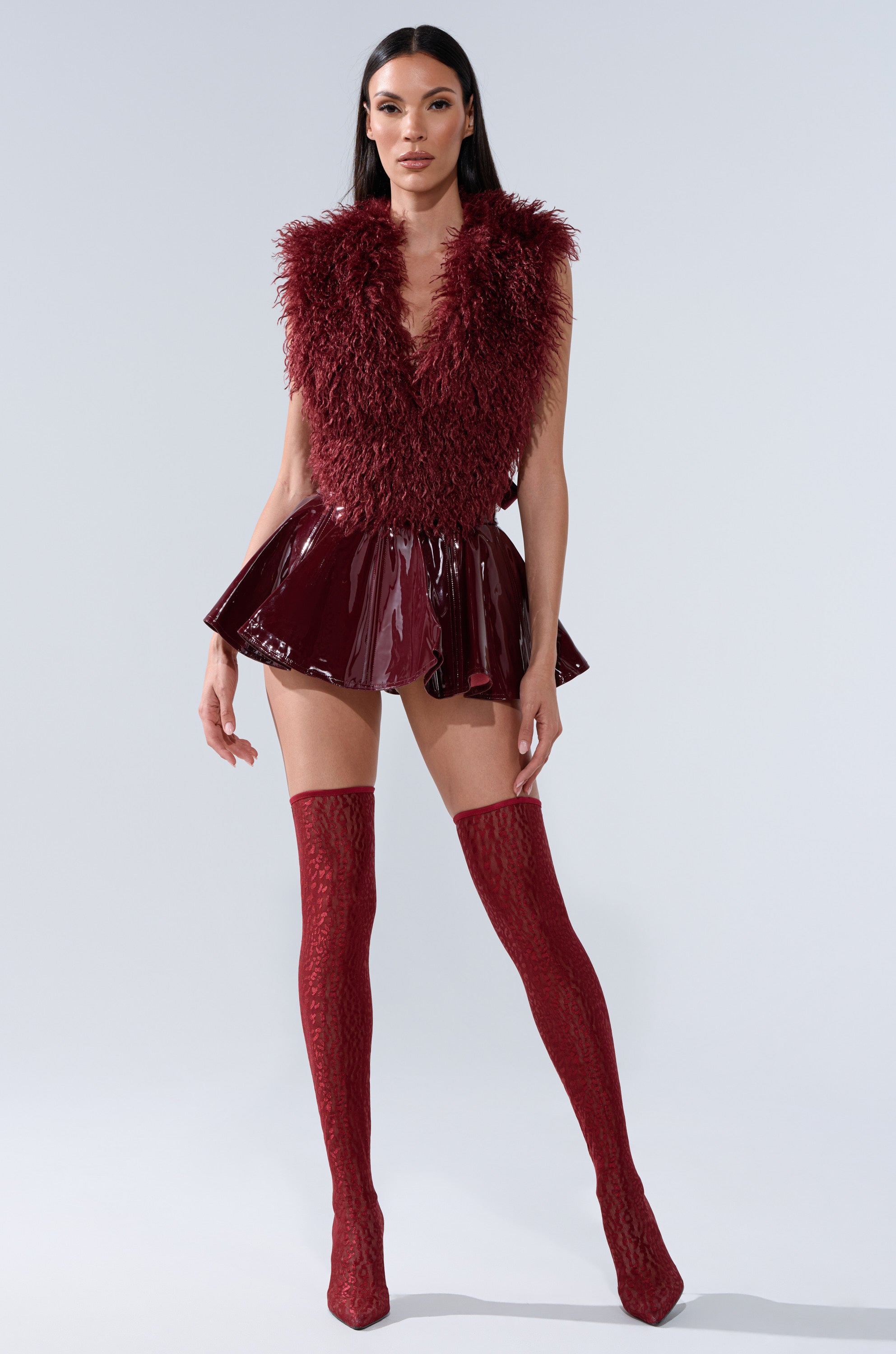 A woman stands confidently in the SHE IS ON FIRE FAUX LEATHER MINI SKIRT IN BURGUNDY, paired with a textured sleeveless top and matching patterned thigh-high boots against a plain light background.