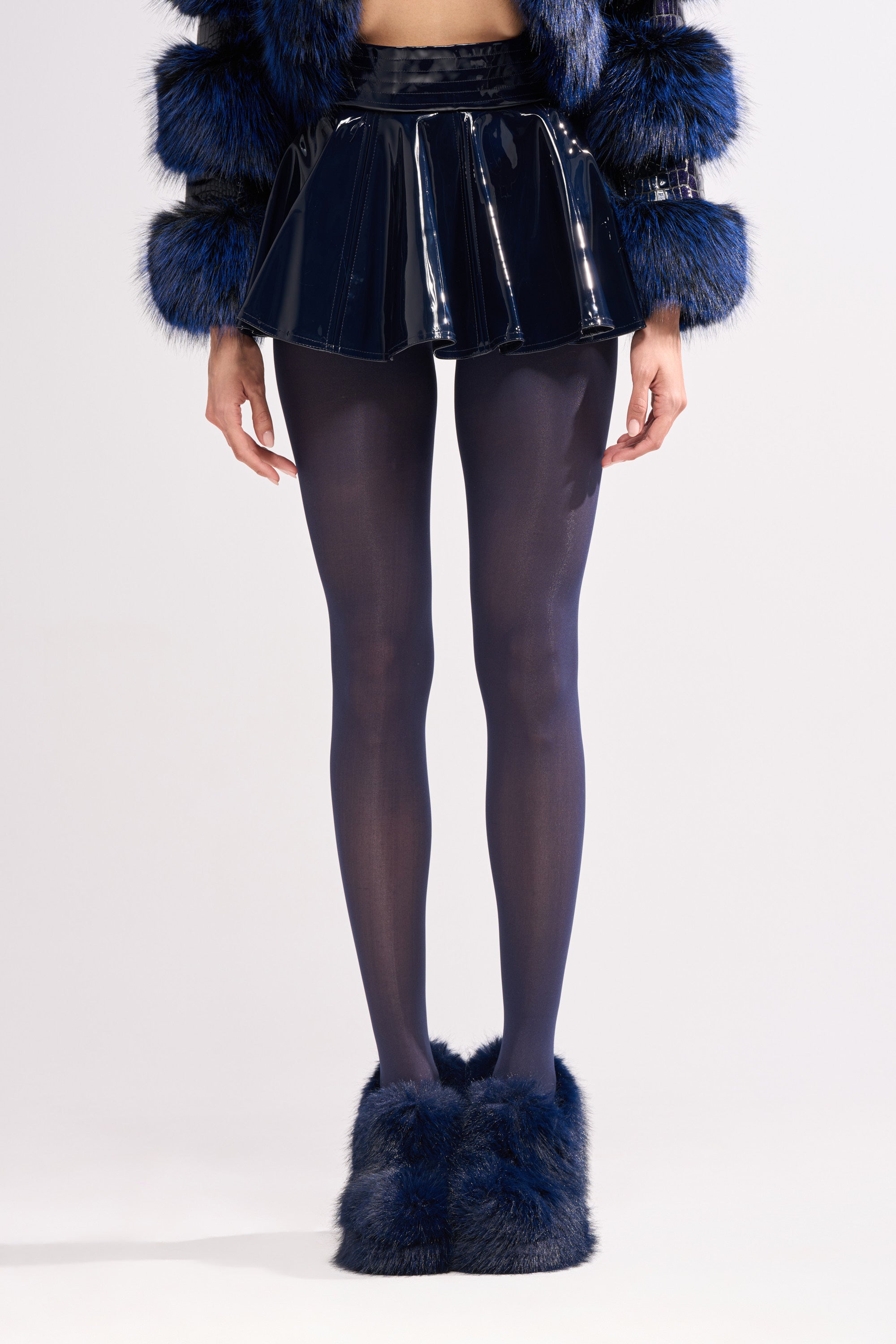 Wearing the SHE IS ON FIRE FAUX LEATHER MINI SKIRT IN NAVY, paired with matching dark blue tights, a fuzzy blue and black jacket, and fluffy blue shoes, a person stands against a plain light background.