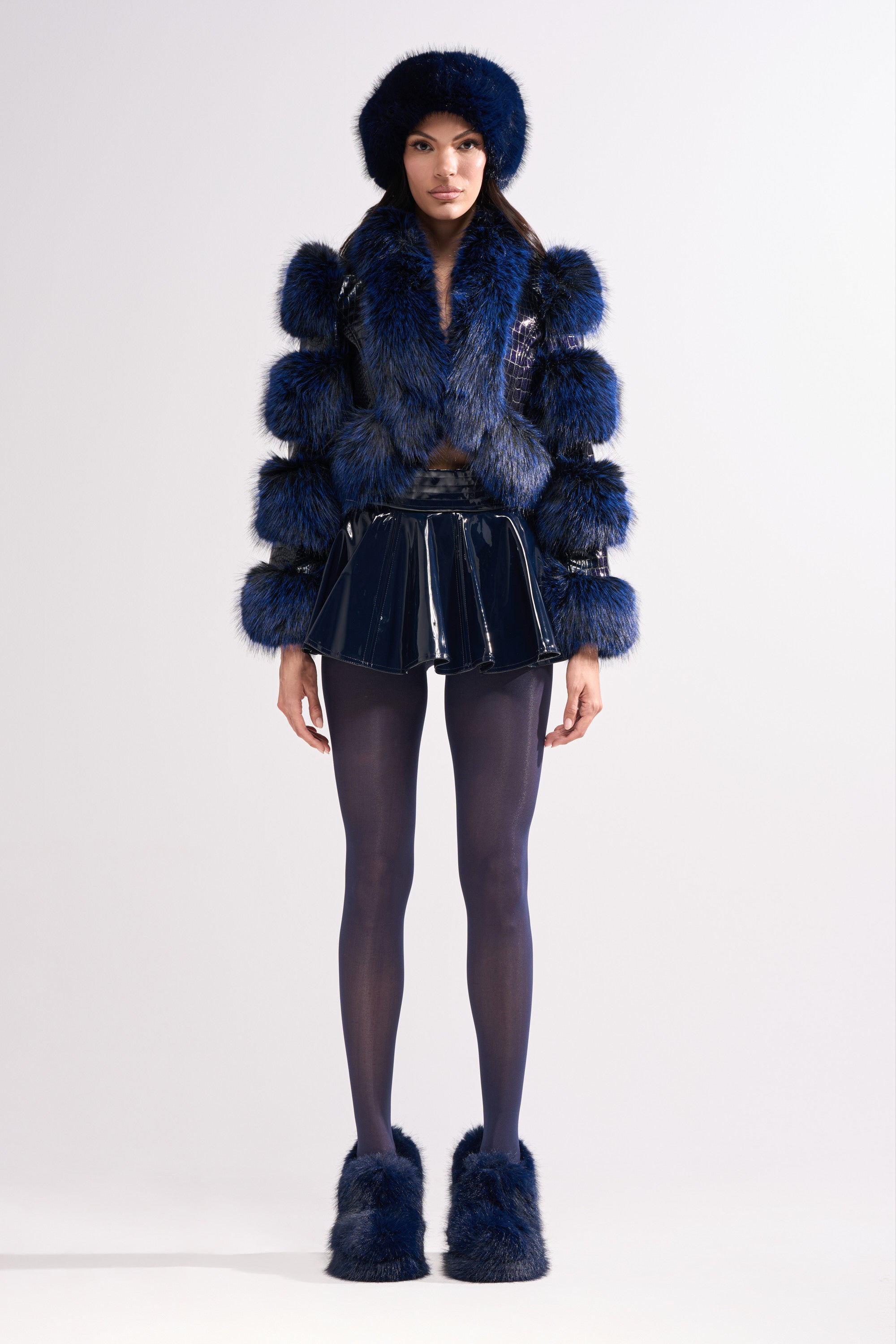 A person wears a dark blue faux-fur hat and jacket with square sections, the SHE IS ON FIRE FAUX LEATHER MINI SKIRT IN NAVY, sheer black tights, and matching furry shoes, posing against a plain light background.
