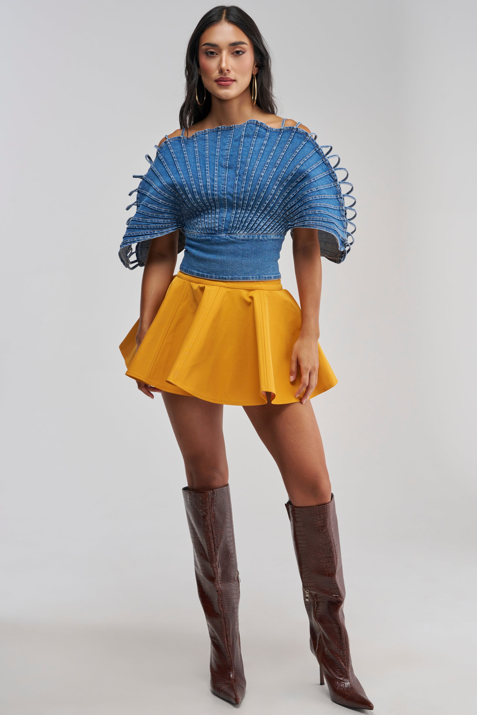 A woman poses against a light gray background wearing the BETTER BE NYLON SKIRT IN MUSTARD, which features a mini length and skater-style hemline. She pairs the skirt with a blue textured top with wide, fan-like sleeves and tall brown boots. Her look is completed with long dark hair and large hoop earrings.