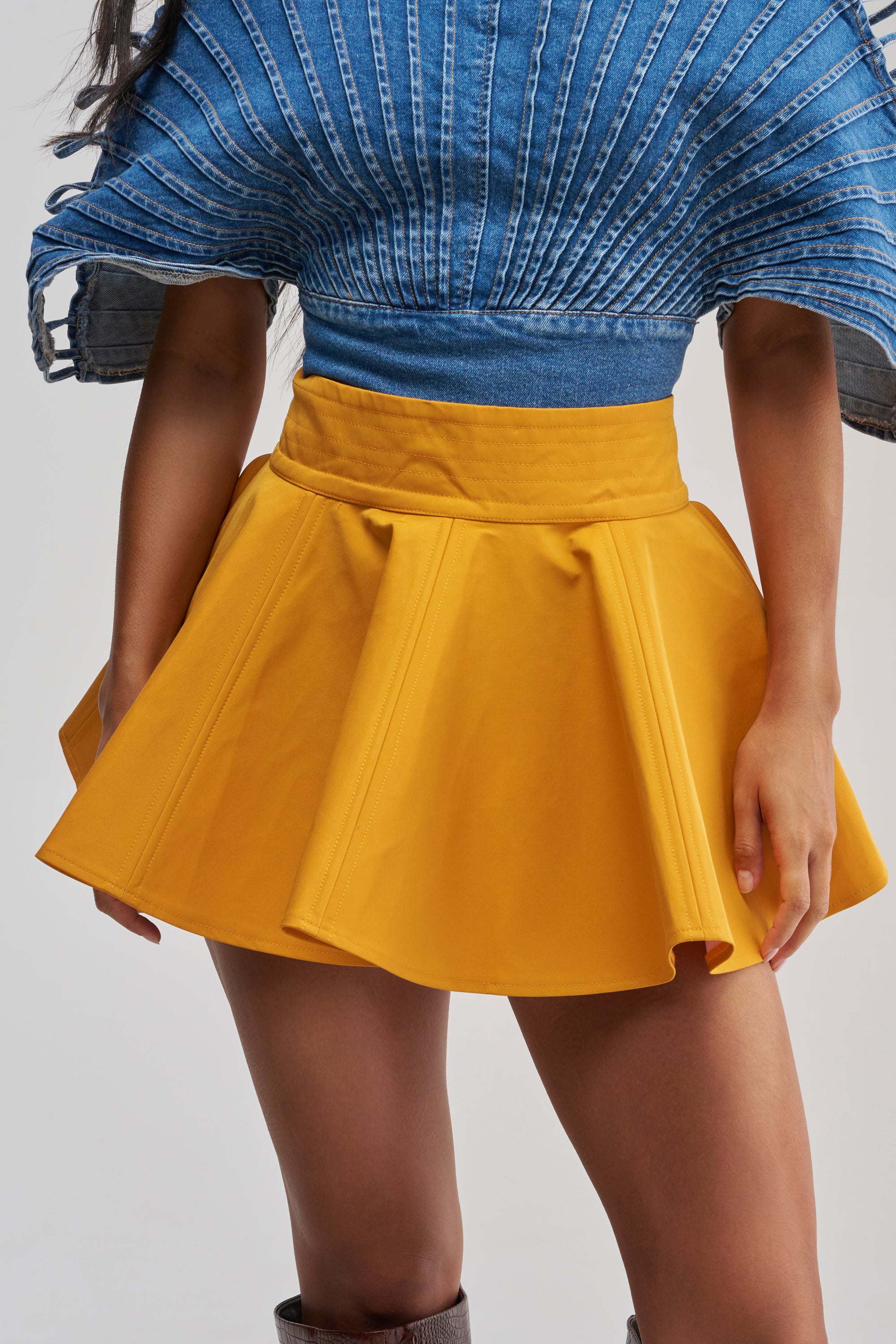 A person wearing a blue textured short-sleeve top paired with the BETTER BE NYLON SKIRT IN MUSTARD, a bright yellow mini skirt with a skater-style hemline, shown from shoulders to mid-thigh.