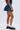 A person wears a dark blue crop top, the BETTER BE DENIM MINI SKIRT with a high-rise, flared skater silhouette, white lace socks, and black chunky loafers while standing against a plain light background.