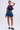 A woman stands against a plain background wearing a navy "FRANCE" crop top, the BETTER BE DENIM MINI SKIRT, white frilly socks, black loafers, and a leopard print handbag on her shoulder.