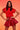 A woman poses confidently against a warm orange gradient, wearing the BETTER BE FAUX LEATHER SKIRT IN BURGUNDY, styled with a shiny red short-sleeved top and gold chain belt. Her hands rest on her hips and her dark hair is loose.