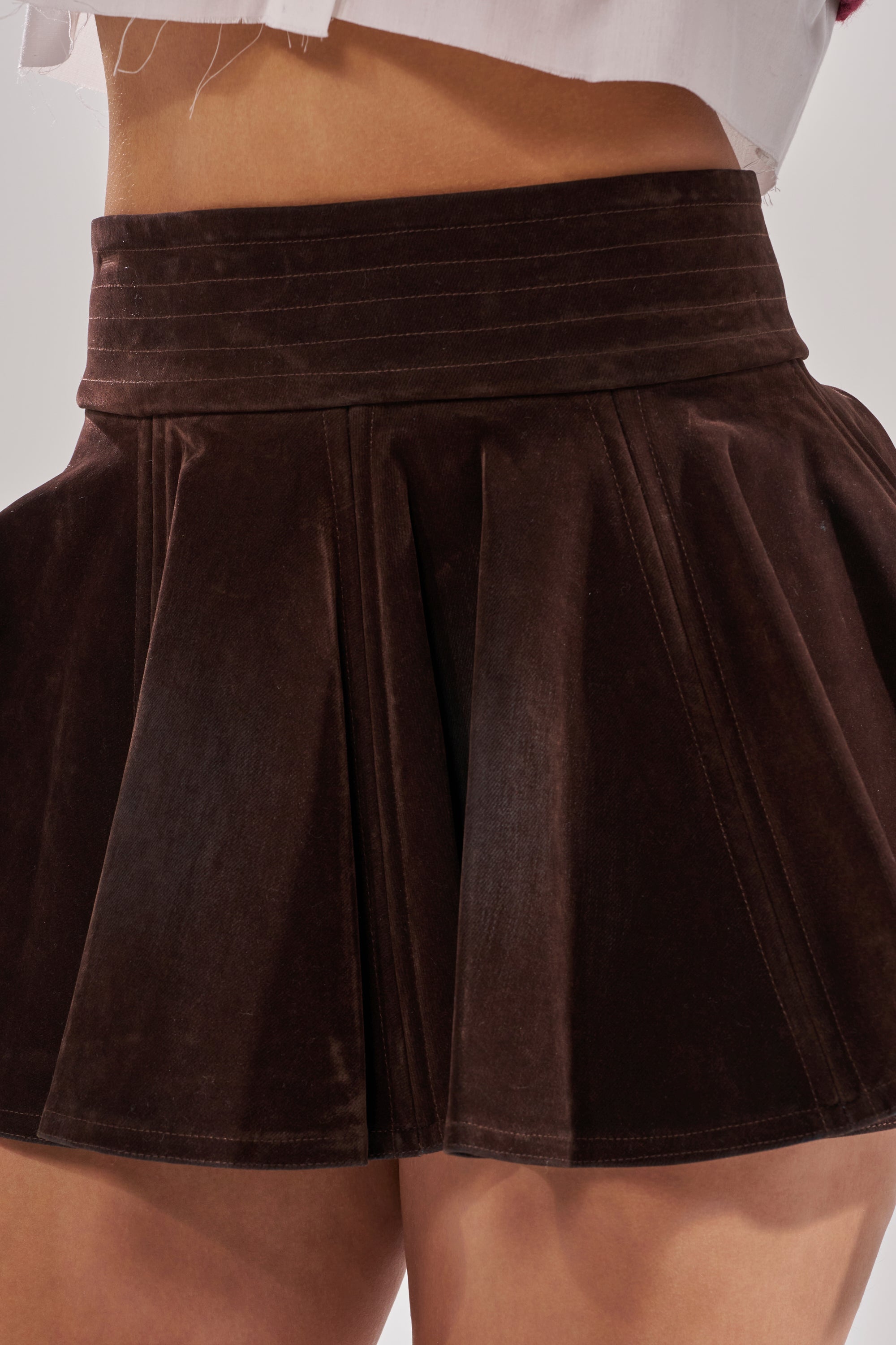 A close-up shows someone wearing the CALL ME VELVET MINI SKIRT in dark brown, featuring pleats and a high waistband. The hem of a white top peeks out above the skirt, highlighting its chic and flattering silhouette.
