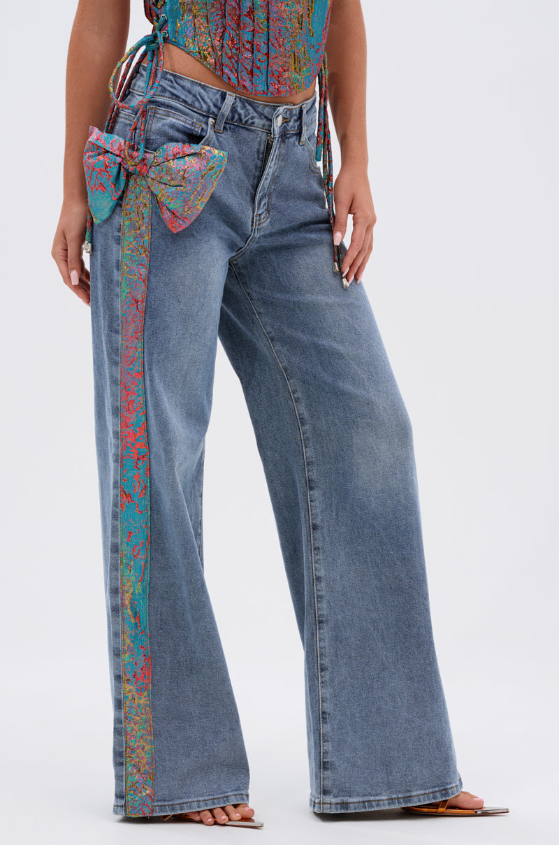 Wearing the PRETTY GIRL PROBLEMS DENIM WITH BOW, these wide-leg blue jeans feature a colorful, patterned vertical stripe and a large matching bow on one side. Only the lower half of the body is shown. 0