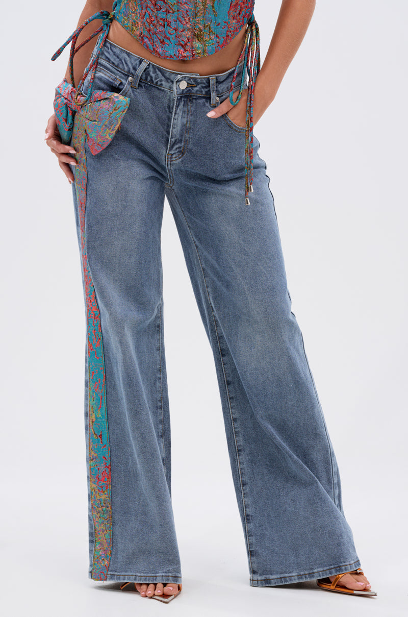 Only the lower half of the body is visible, showcasing PRETTY GIRL PROBLEMS DENIM WITH BOW: wide-leg blue jeans featuring a colorful patterned stripe down one side, styled with a matching crop top and small handbag. 1