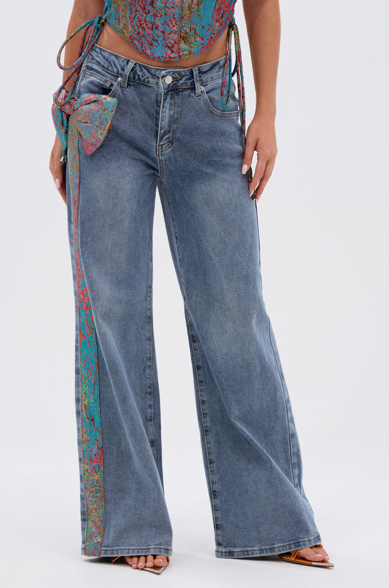 Someone stands against a plain light background wearing the PRETTY GIRL PROBLEMS DENIM WITH BOW—wide-leg jeans featuring a colorful patterned strip and metallic bow—paired with a vibrant matching top and open-toe heels. 2