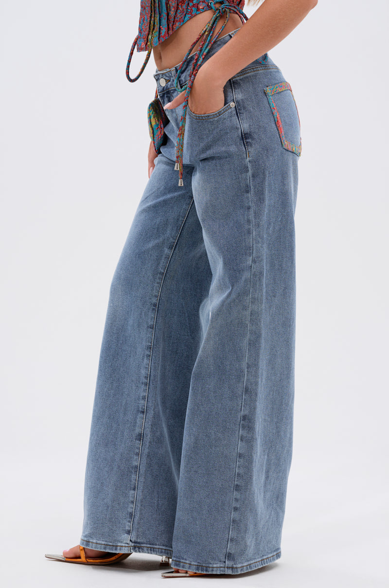 A person wears PRETTY GIRL PROBLEMS DENIM WITH BOW high-waisted, wide-leg jeans with colorful pocket trim, styled with open-toe sandals and a patterned crop top. The side view emphasizes the jeans' loose fit. 3