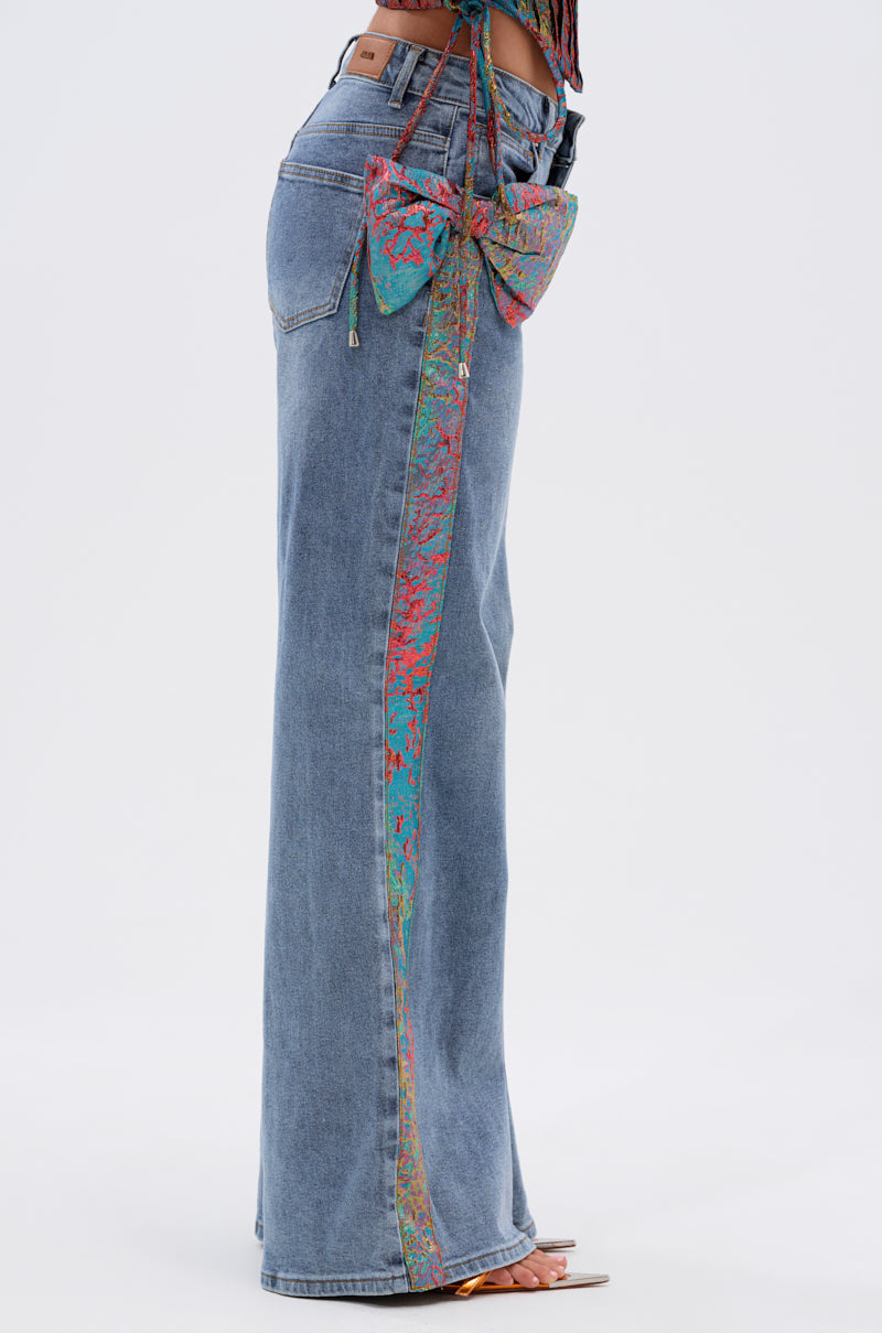 A person wears PRETTY GIRL PROBLEMS DENIM WITH BOW—high-waisted, wide-leg jeans with a vibrant metallic bow appliqué and matching side stripe—paired with open-toe heels, set against a plain white background. 4