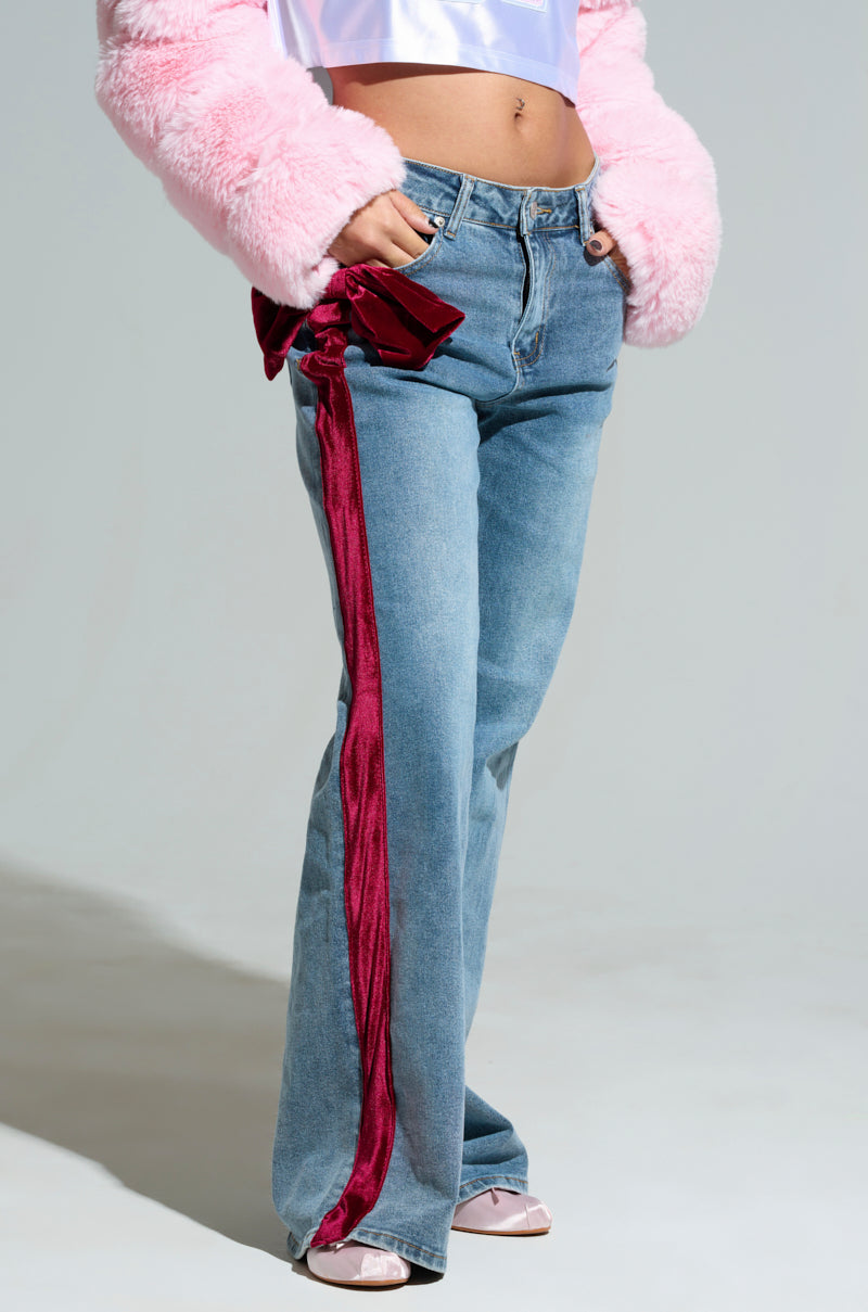 A person showcases the UNWRAP ME VELVET BOW DENIM PANT, featuring a striking red velvet stripe and bow along the side. These wide-leg jeans are styled with a pink furry jacket and coordinating pink shoes, all set against a simple gray backdrop.
