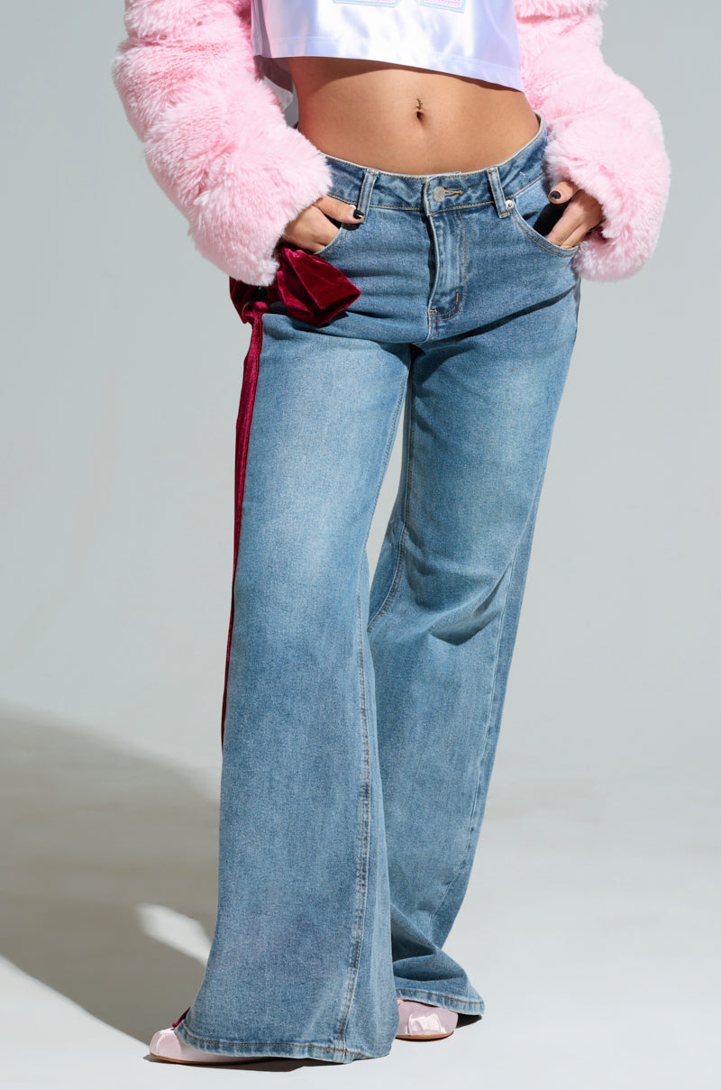 A person wearing UNWRAP ME VELVET BOW DENIM PANTS in light blue with a wide-leg silhouette and a red stripe on the side, paired with a white crop top and a pink fuzzy jacket. Their hands are tucked into the pockets of the high-rise denim.