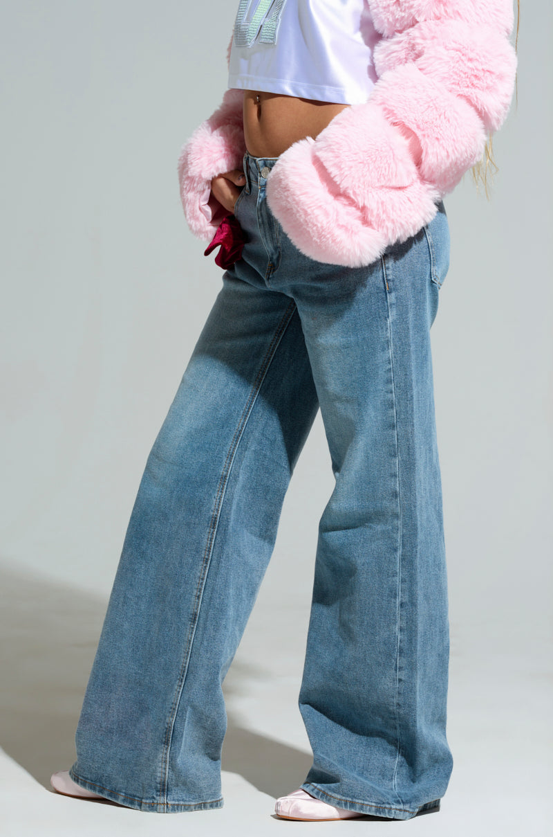 A person is wearing the UNWRAP ME VELVET BOW DENIM PANT with a white T-shirt and a pink fluffy jacket. Only the lower half of the body is visible, highlighting the denim pants and accessories.
