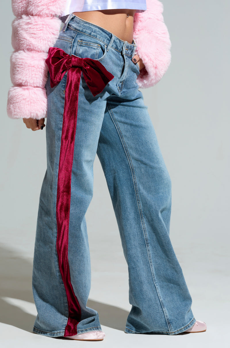 A person is wearing the UNWRAP ME VELVET BOW DENIM PANT, which features wide-leg denim jeans with a red velvet stripe and a velvet bow on the side. They're paired with a pink fuzzy jacket that shows part of their midriff and light pink shoes against a plain background.