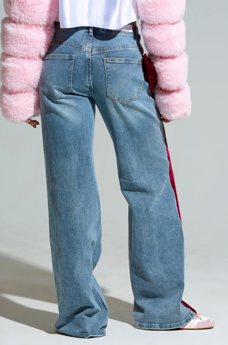 A person wearing the Unwrap Me Velvet Bow Denim Pant, featuring high-rise denim with a wide-leg silhouette and a red stripe along the side, stands with their back to the camera. They pair it with a pink fluffy jacket and pale pink shoes. The background is plain and neutral.