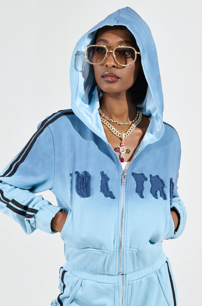 Dressed in a light blue "DON'T TALK TO ME I'M RUNNING ERRANDS" zip-up sweatshirt and matching pants, a person strikes a pose with oversized sunglasses, a gold chain necklace, and an ombré multicolored pendant. Their cropped silhouette stands out against the plain white background.