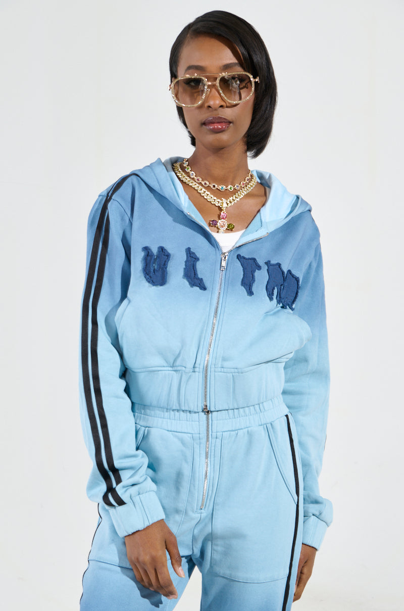 The person is dressed in the DON'T TALK TO ME I'M RUNNING ERRANDS ZIP UP SWEATSHIRT, which features a fashionable blue color with black stripes and distinct design elements, including an ombré effect. They enhance their look with sunglasses and layered necklaces, set against a plain, light-colored background.