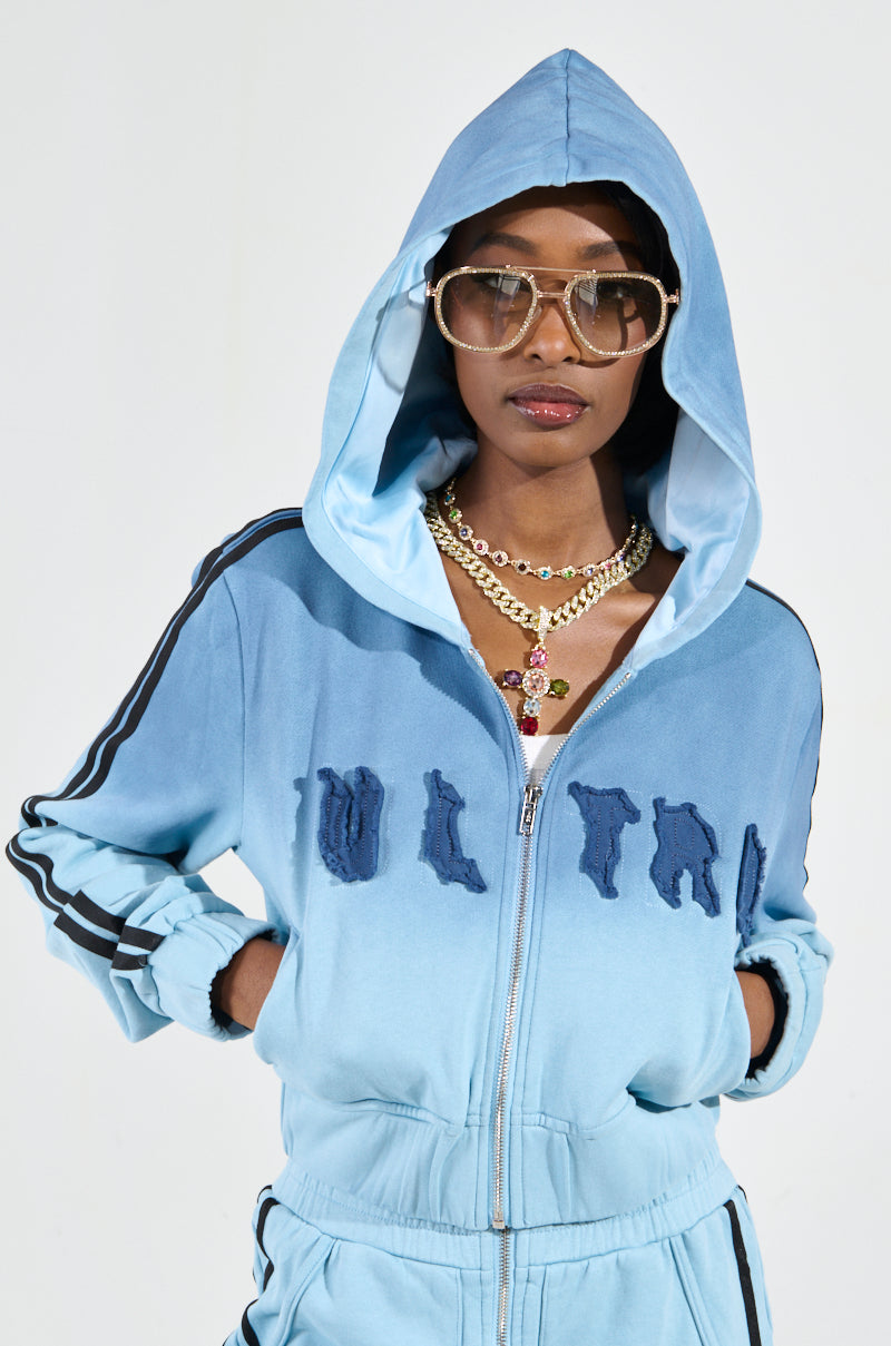 Dressed in the chic, ombré blue "DON'T TALK TO ME I'M RUNNING ERRANDS" zip-up sweatshirt with a stylish cropped silhouette, a person exudes fashion-forward elegance. Their look is completed with large aviator sunglasses and layered gold necklaces featuring a pendant, all set against a simple background that accentuates their ensemble.